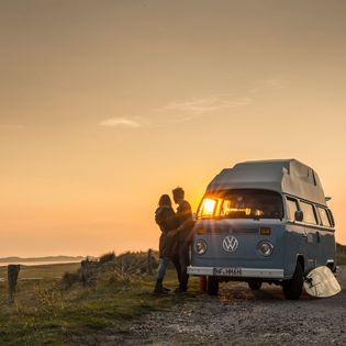 Paar lehnt an VW-Bulli bei Sonnenuntergang am Ellenbogen.