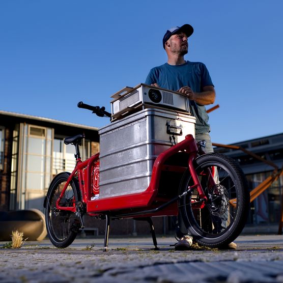 Mann steht mit rotem Lastenrad mit Projektor vor einem Gebäude.