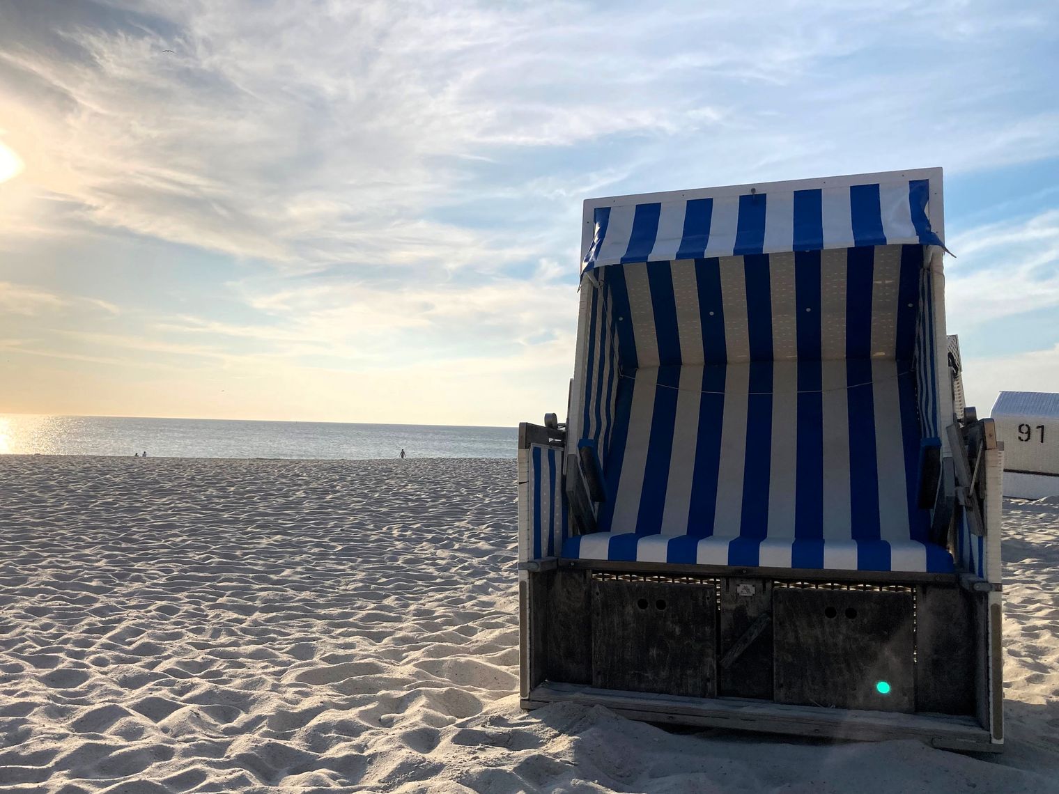 Ein leerer blau-weiß gestreifter Strandkorb am hellen Sandstrand mit Blick auf die ruhige Nordsee bei Sonnenuntergang.