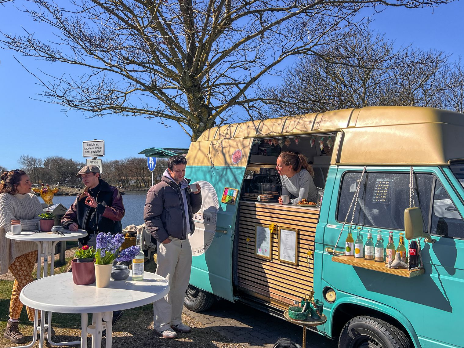 ein restaurierter VW T3 Bus steht vor dem Dorfteich. Einige Personen stehen an stehtischen davor.