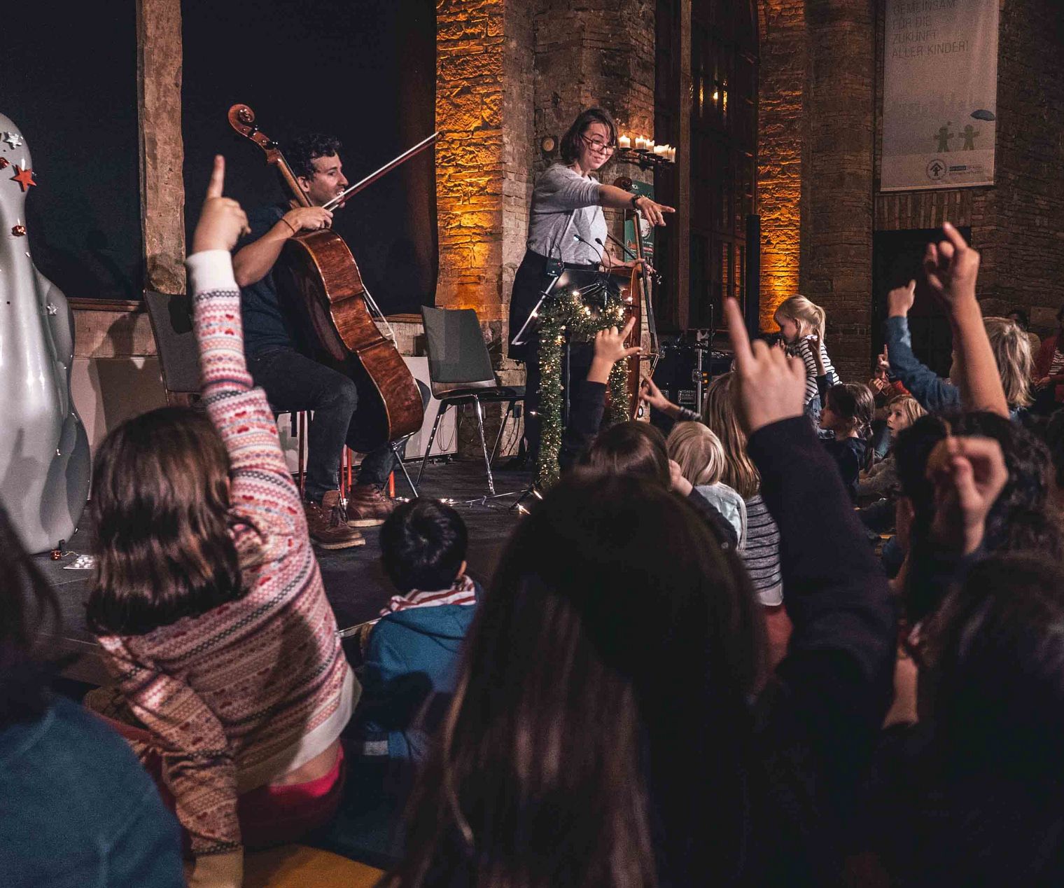 Kinder heben die Hände bei einem interaktiven Konzert mit Cello und Moderatorin in stimmungsvoller Kulisse.