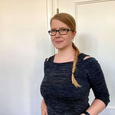 Frau mit Brille und Zopf steht vor weißer Tür und lächelt.