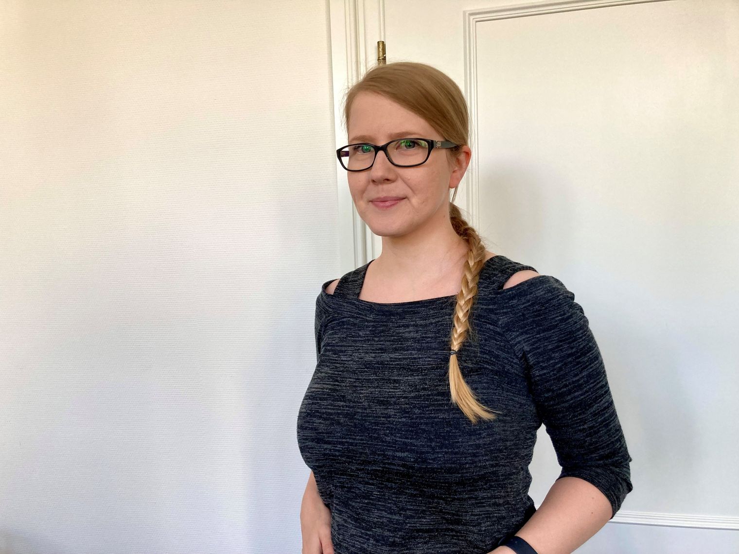 Frau mit Brille und Zopf steht vor weißer Tür und lächelt.
