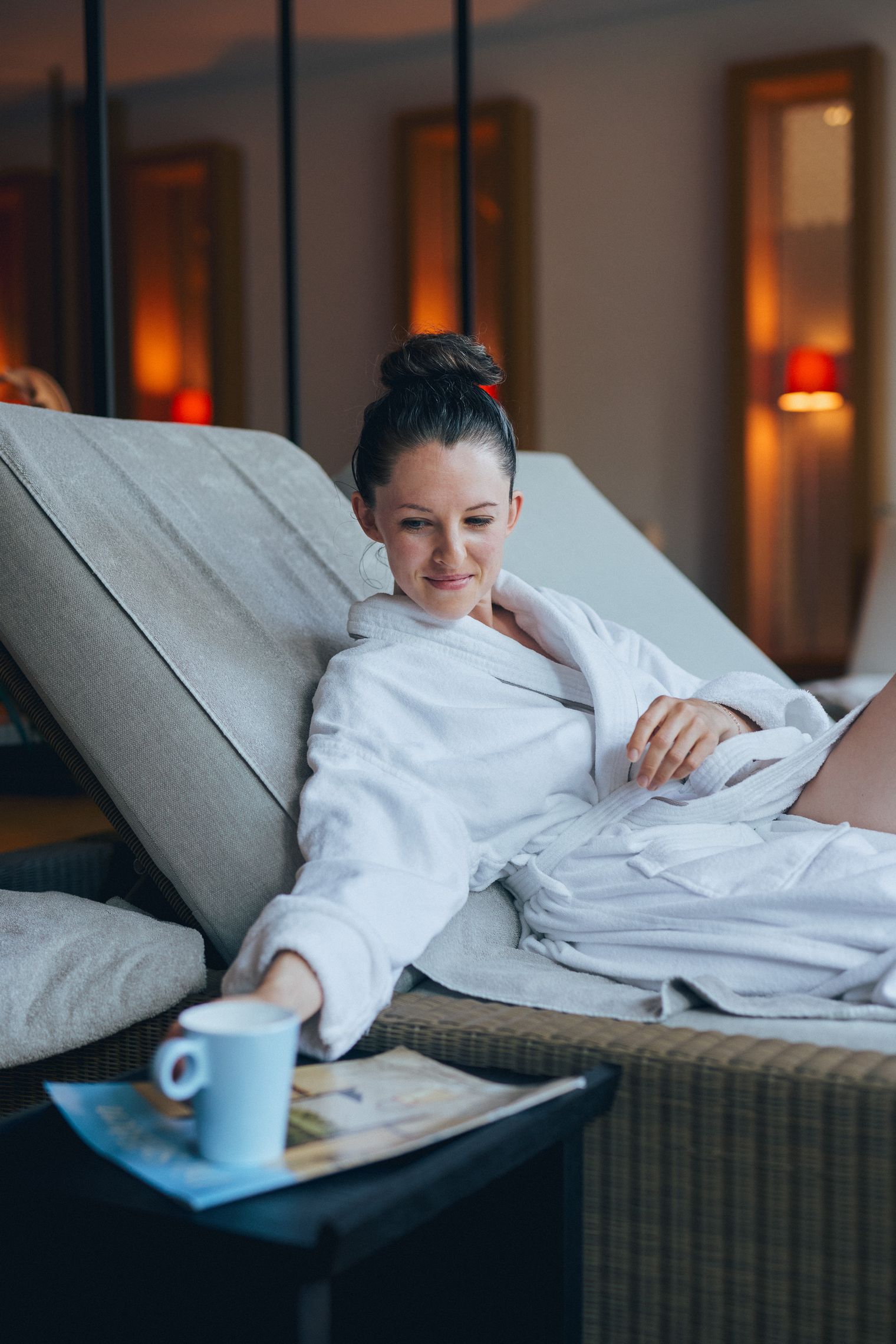 Eine Frau im weißen Bademantel liegt entspannt auf einer Liege und greift nach einer blauen Tasse auf einem kleinen Tisch neben ihr.
