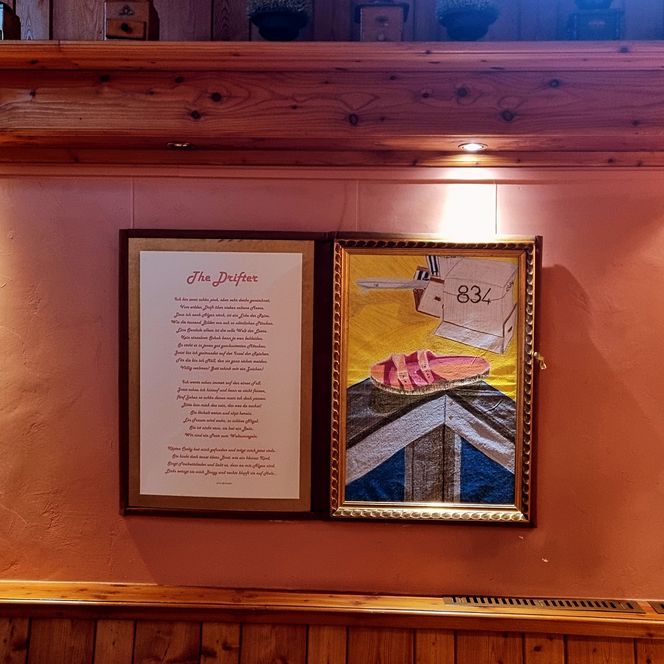 Zwei gerahmte Bilder an einer Holzwand, links ein Gedicht, rechts ein Bild mit Sandale und Strandmotiv.