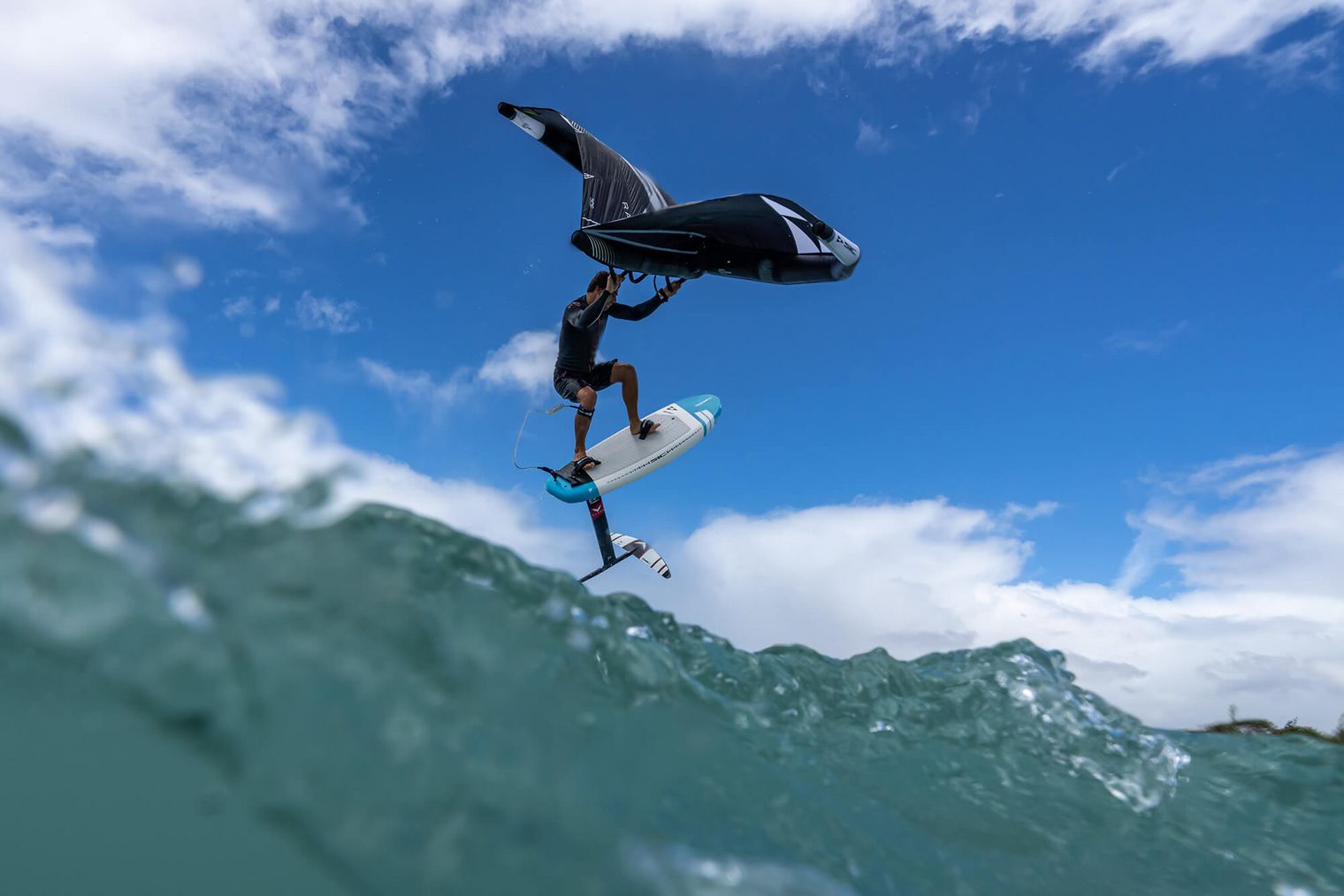 Ein Wingfoiler auf einem Foilboard hebt über den Wellen ab, mit einem strahlend blauen Himmel und weißen Wolken im Hintergrund.