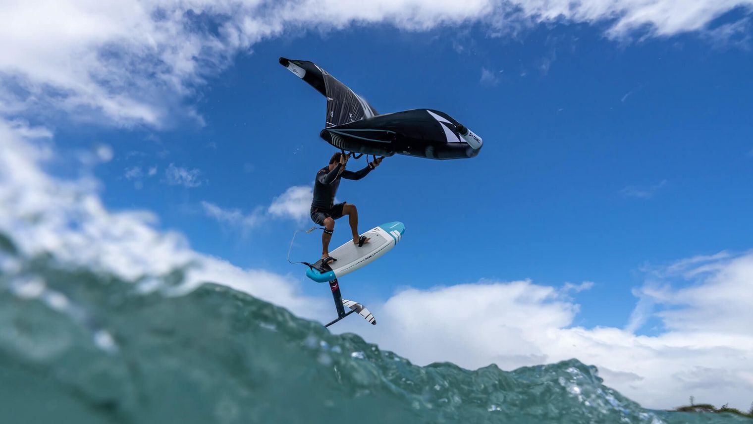 Ein Wingfoiler auf einem Foilboard hebt über den Wellen ab, mit einem strahlend blauen Himmel und weißen Wolken im Hintergrund.