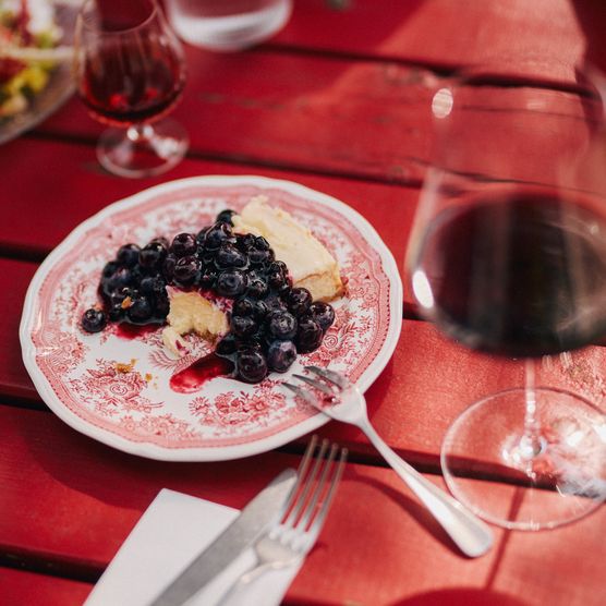 Käsekuchen mit Blaubeeren auf einem roten Holztisch im Käseklub in Keitum auf Sylt, mit Rotwein und Wasser im Hintergrund. Bildunterschrift: Genuss im Käseklub in Keitum auf Sylt – hausgemachter Käsekuchen mit frischen Blaubeeren.
