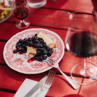 Käsekuchen mit Blaubeeren auf einem roten Holztisch im Käseklub in Keitum auf Sylt, mit Rotwein und Wasser im Hintergrund. Bildunterschrift: Genuss im Käseklub in Keitum auf Sylt – hausgemachter Käsekuchen mit frischen Blaubeeren.