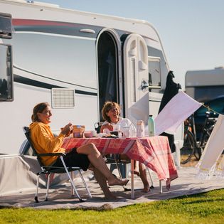 Zwei Frauen frühstücken und lachen vor ihrem Wohnwagen.
