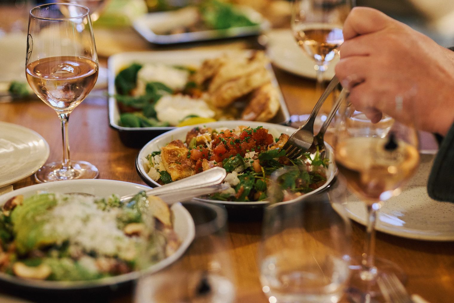 Verschiedene mediterrane Speisen und Gläser Roséwein auf einem gedeckten Tisch, eine Person nimmt eine Gabel voll Essen.