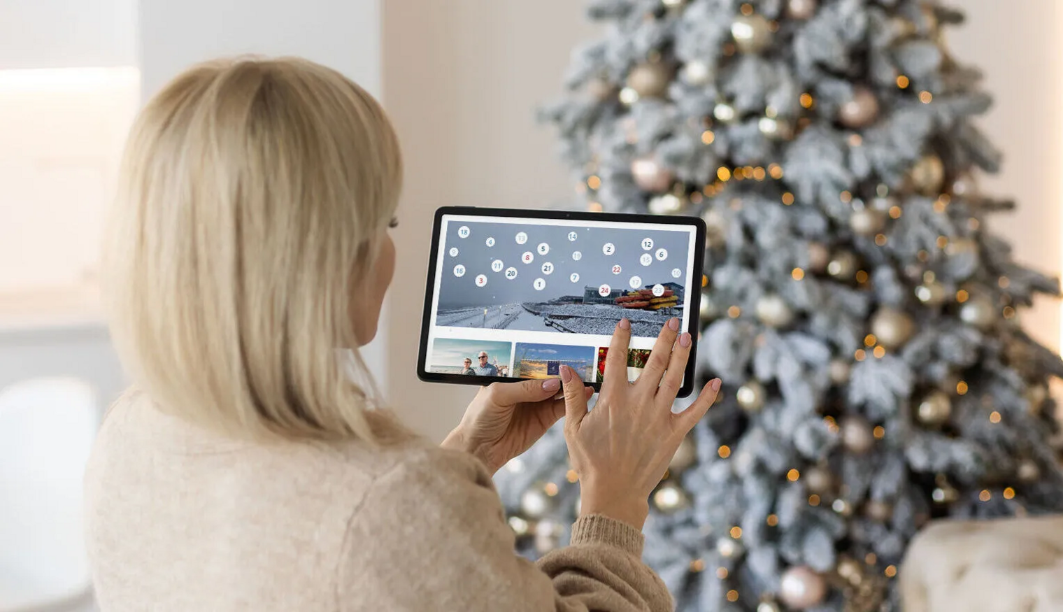 Frau tippt auf digitalen Adventskalender auf ihrem Tablet - im Hintergrund leuchtet ein geschmückter Tannenbaum