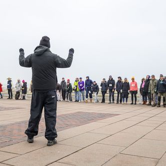Eine Person mit erhobenen Armen steht vor einer Reihe warm gekleideter Teilnehmender auf einer windigen Strandpromenade.