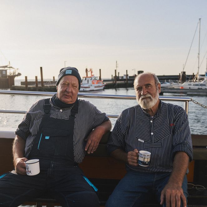 Zwei ältere Männer in gestreiften Hemden sitzen entspannt auf einem Boot im Hafen und halten Kaffeetassen in den Händen.