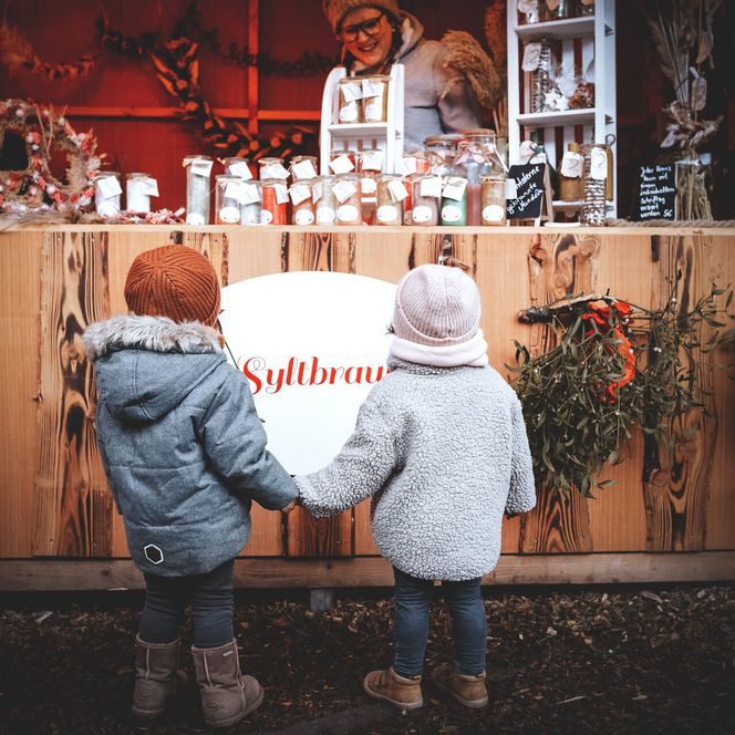 Zwei Kinder stehen Hand in Hand vor einem weihnachtlichen Marktstand.