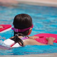 Ein Kind mit Schwimmbrille und Schwimmhilfe auf dem Rücken gleitet mit einem roten Schwimmbrett durch das Wasser.
