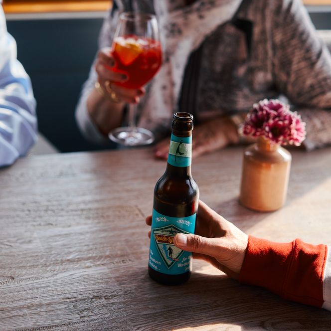 Drei Personen sitzen an einem Tisch mit Getränken. Eine hält eine Bierflasche, die anderen Cocktails. Eine Vase mit rosa Blume steht auf dem Tisch.
