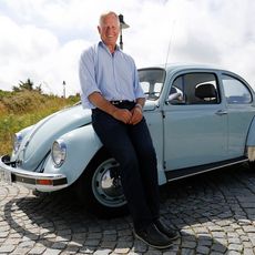 Mann sitzt auf einem blauen VW Käfer bei Sonnenschein