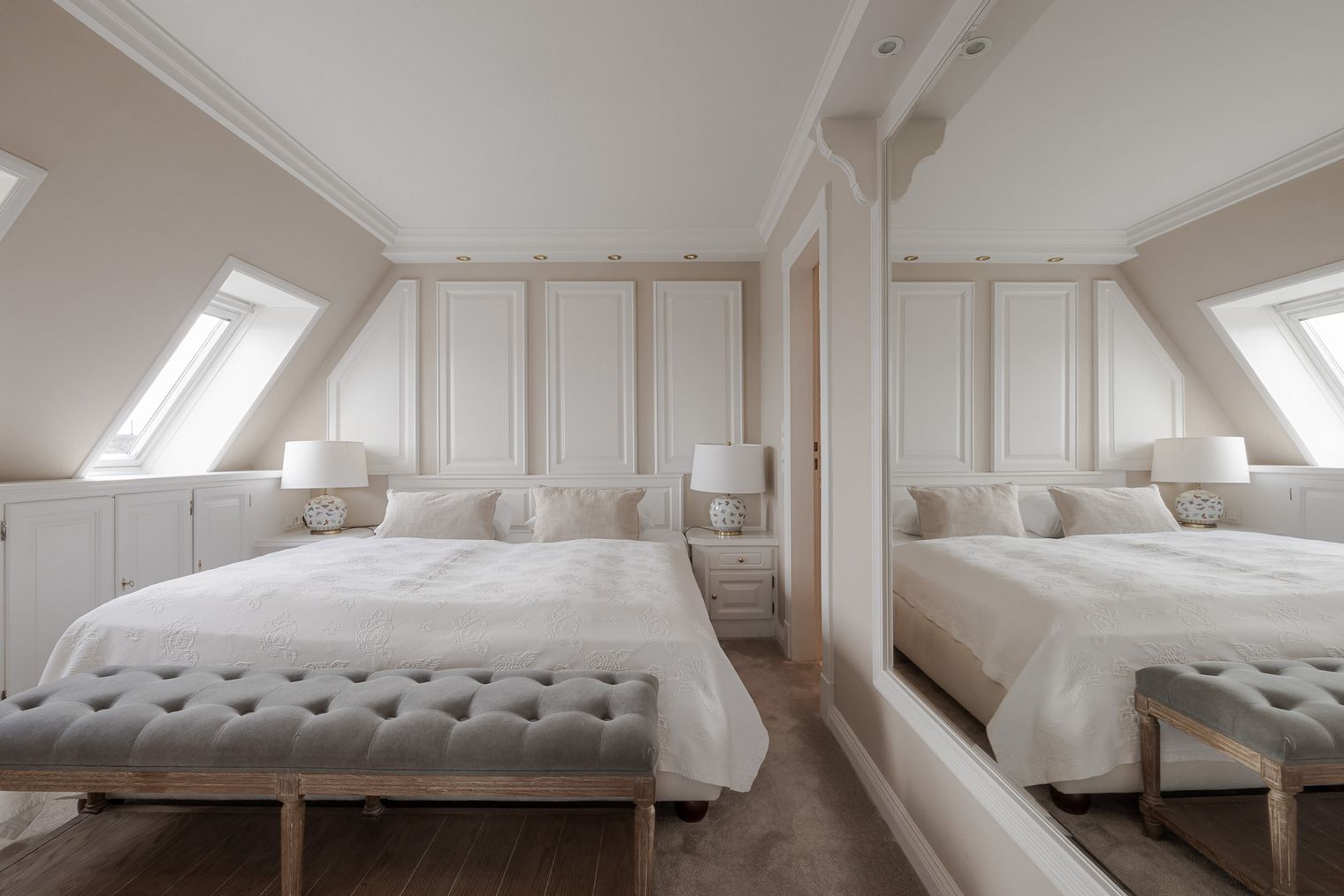 Helles Schlafzimmer mit weißem Doppelbett, grauer Sitzbank, Wandpaneelen, Dachfenstern und großem Spiegel.