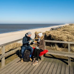 Ein Paar trinkt aus einer Thermoskanne auf einer Holzbank mit Blick auf den Kampener Strand