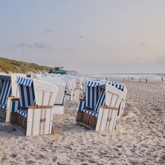 Reihe von weiß-blauen Strandkörben am Sandstrand, mit Blick auf das Meer und sanft beleuchtete Dünen im Hintergrund. Einige Personen spazieren am Wasser entlang.