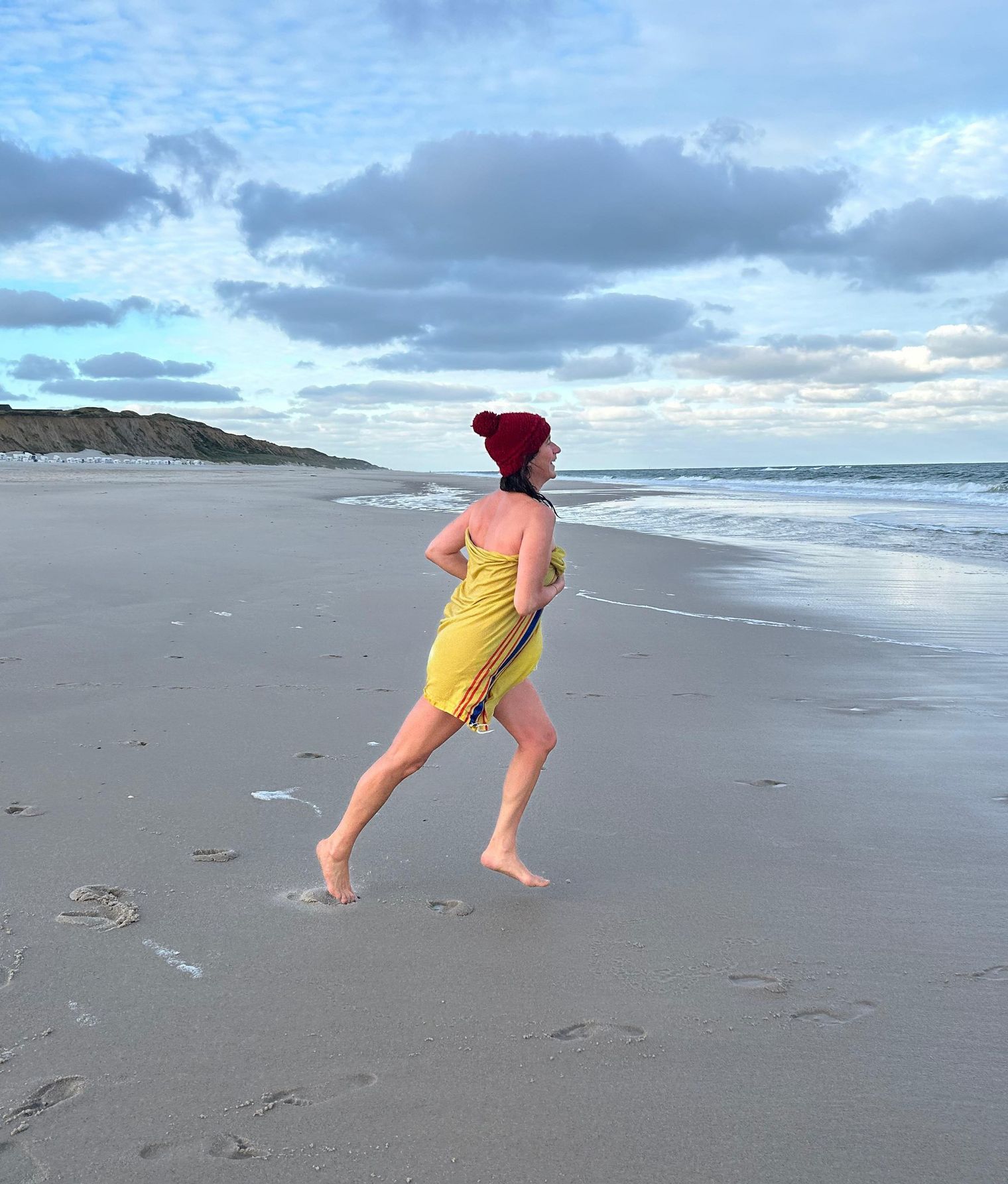 Person mit roter Mütze läuft barfuß im Handtuch am Strand.