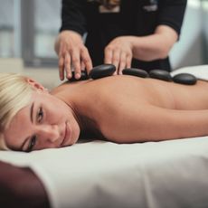 Eine blonde Frau erhält eine Hot-Stone-Massage auf einer Massageliege in einem hellen Wellnessraum.