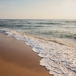 Foamy waves hit a calm sandy beach in light sunlight.