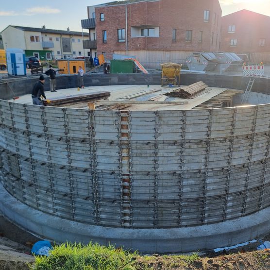 Bauarbeiter montieren Schalung und Holzplatten auf rundem Eisspeicher im Neubaugebiet.