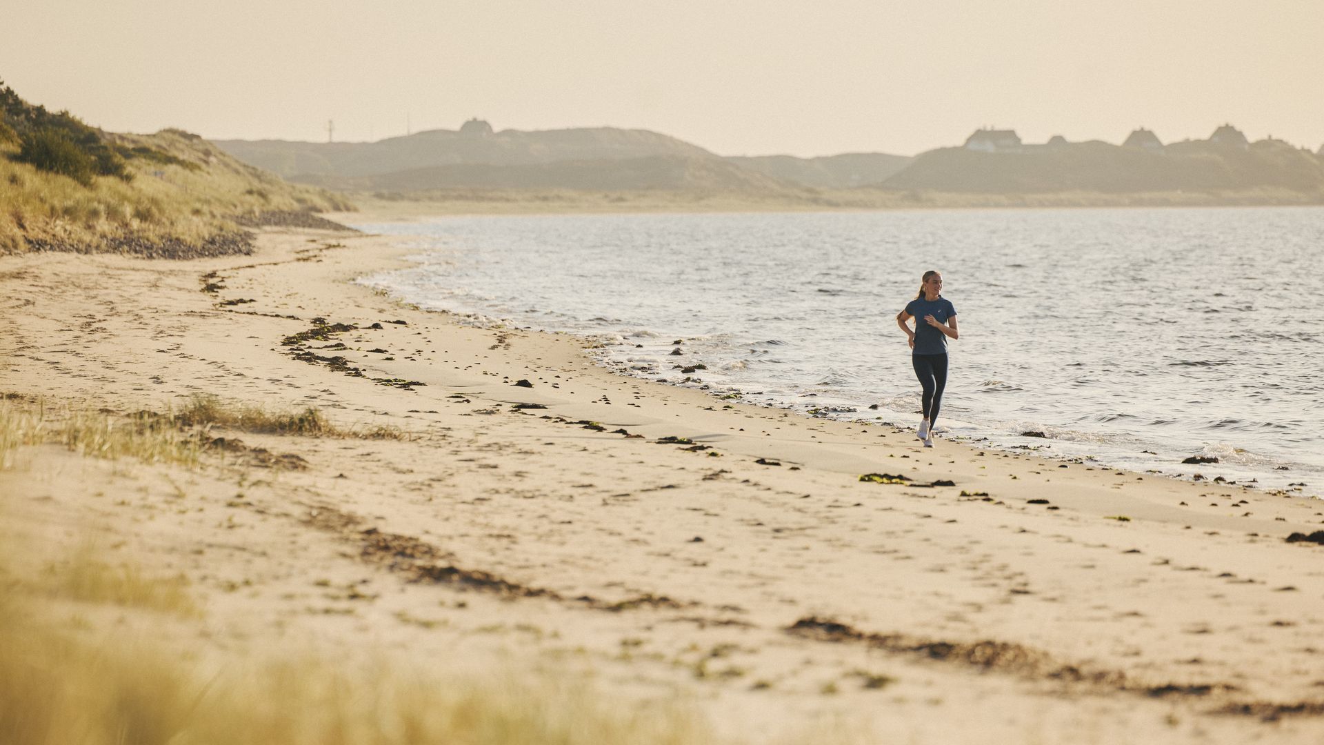 Eine Frau läuft am Sandstrand entlang, die Weite des Meeres und die sanften Hügel der Dünenlandschaft bilden eine ruhige Kulisse.