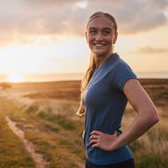 Eine Frau lächelt mit Händen in der Hüfte auf einem Pfad, umgeben von Wiesen und Sonnenaufgang im Hintergrund.