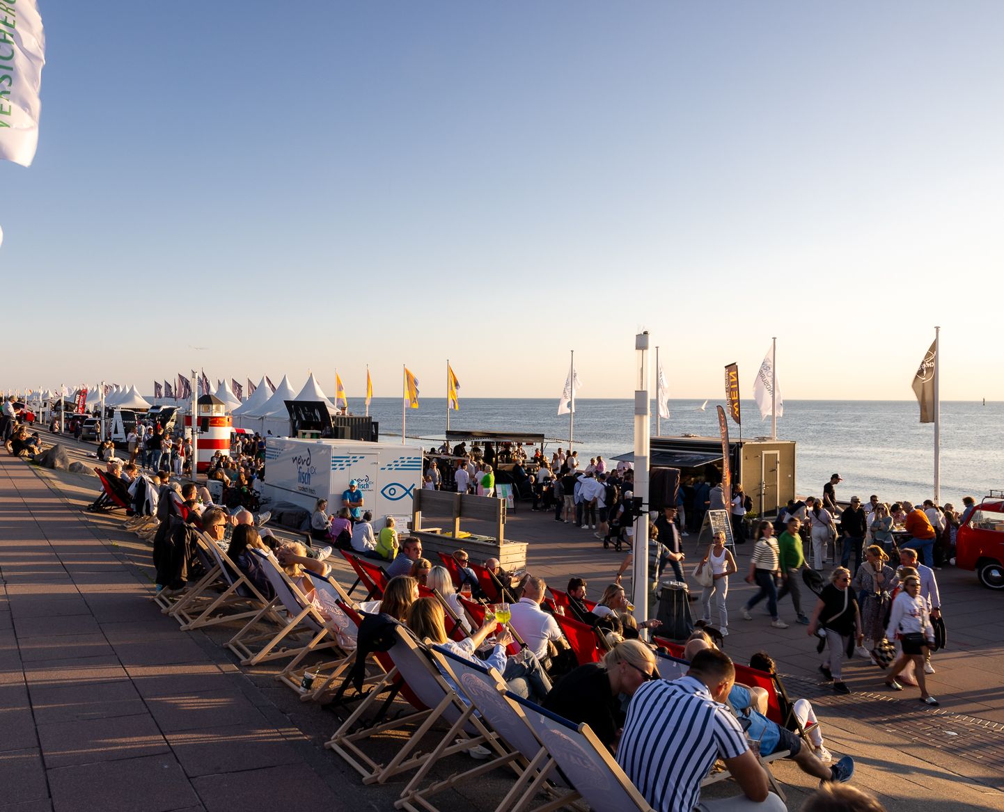 Belebte Westerländer Strandpromenade mit Besuchern in Liegestühlen, Veranstaltungszelten, Foodtrucks und bunten Flaggen am Meer im warmen Abendlicht.