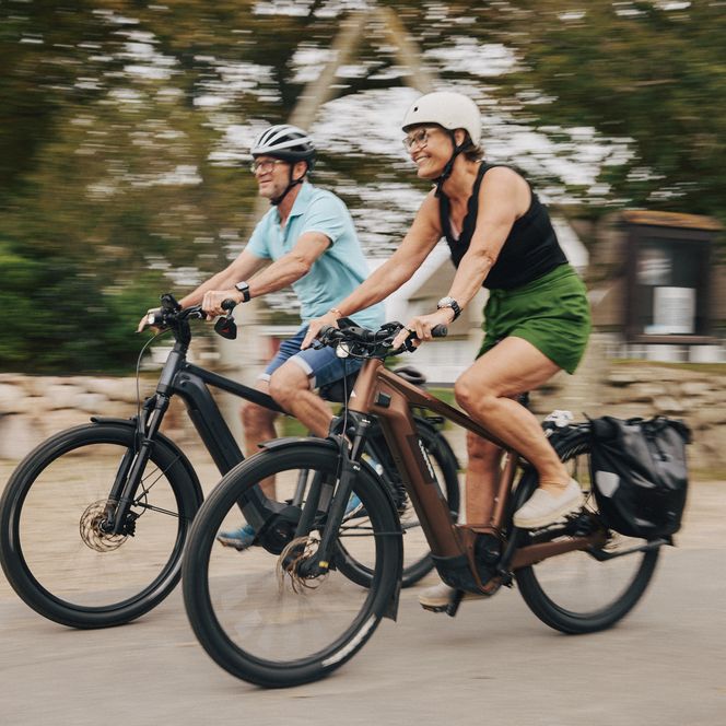 Zwei Personen auf E-Bikes fahren durch Keitum auf Sylt. Im Hintergrund sind Reetdachhäuser sichtbar.